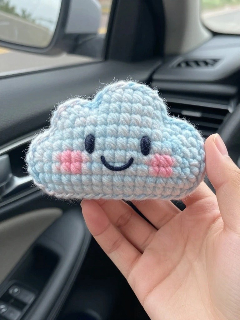 A light blue crocheted cloud with a smiling face and pink blush cheeks clips onto a car air vent, held by a hand inside a vehicle.