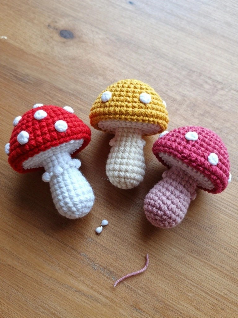Three small handmade crocheted mushrooms in red, yellow, and pink with white-spotted caps and stems on a wooden table next to yarn scraps.