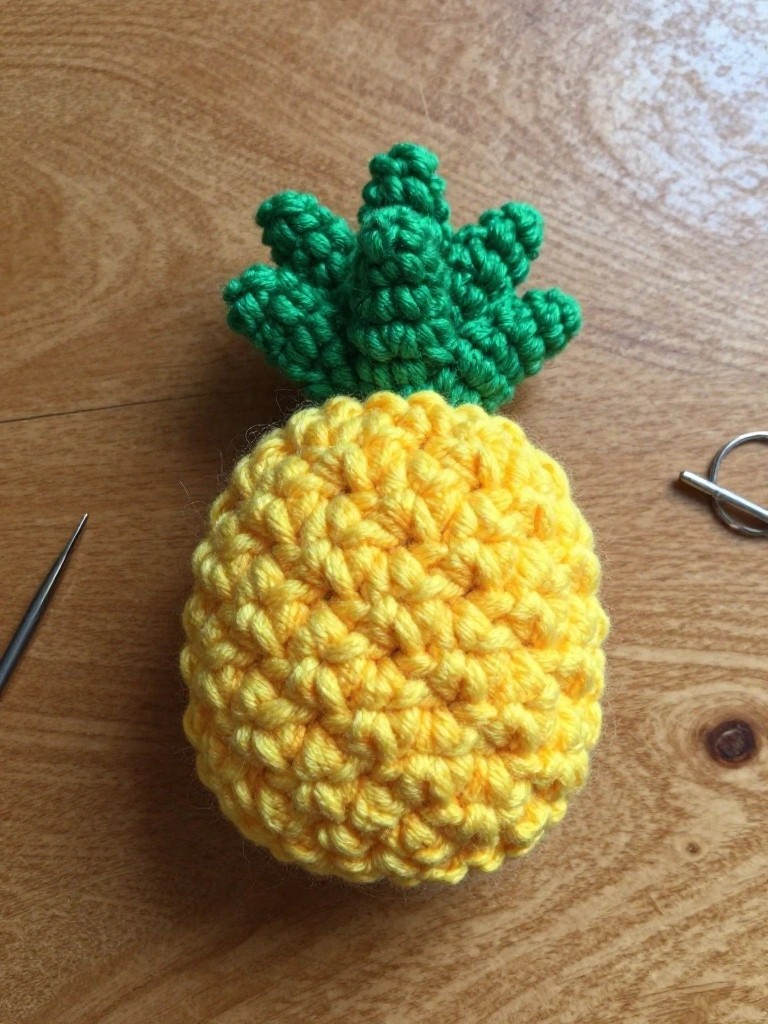 A small yellow crocheted pineapple with green top leaves sits on a wooden table next to a silver crochet hook and metal ring.
