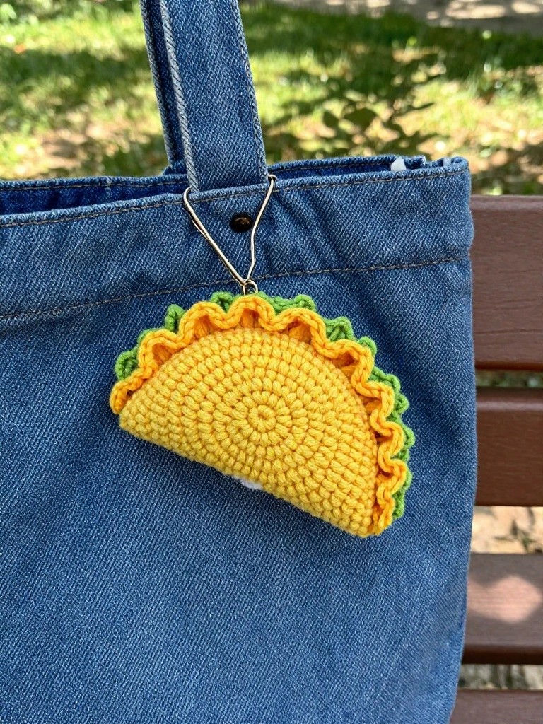 Crocheted yellow taco charm with green ruffled edges hanging from the strap of a blue denim tote bag on an outdoor bench.