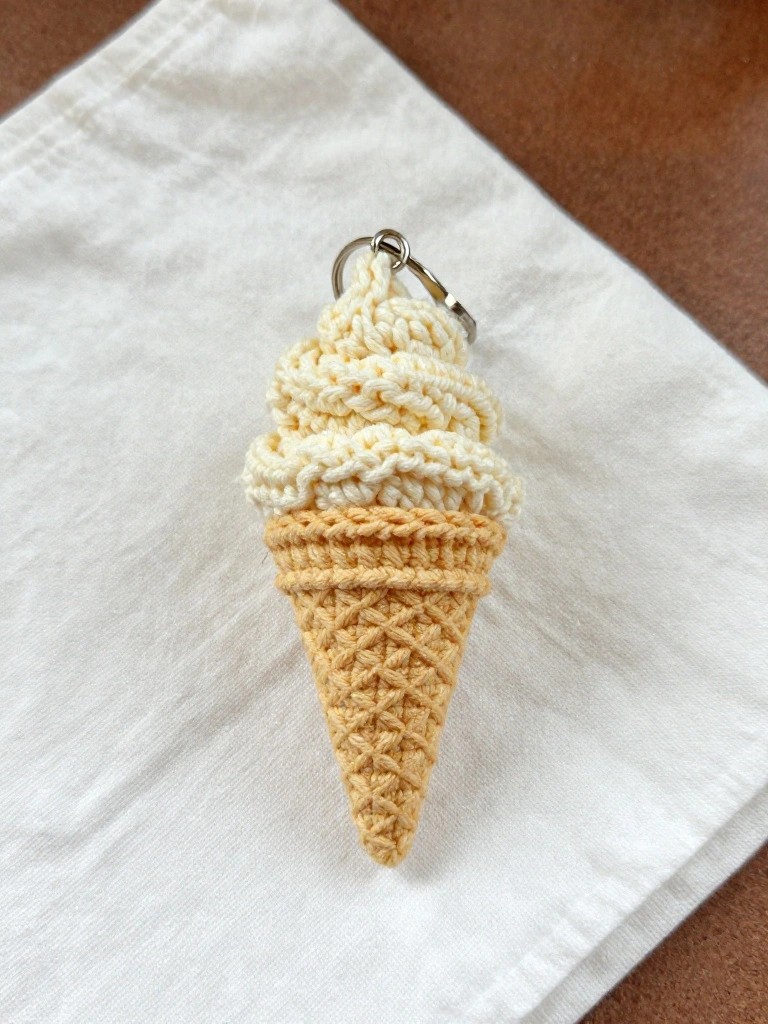 A handmade crochet keychain shaped like a vanilla ice cream cone in light tan and creamy white yarn sits on white fabric with a silver keyring attached.