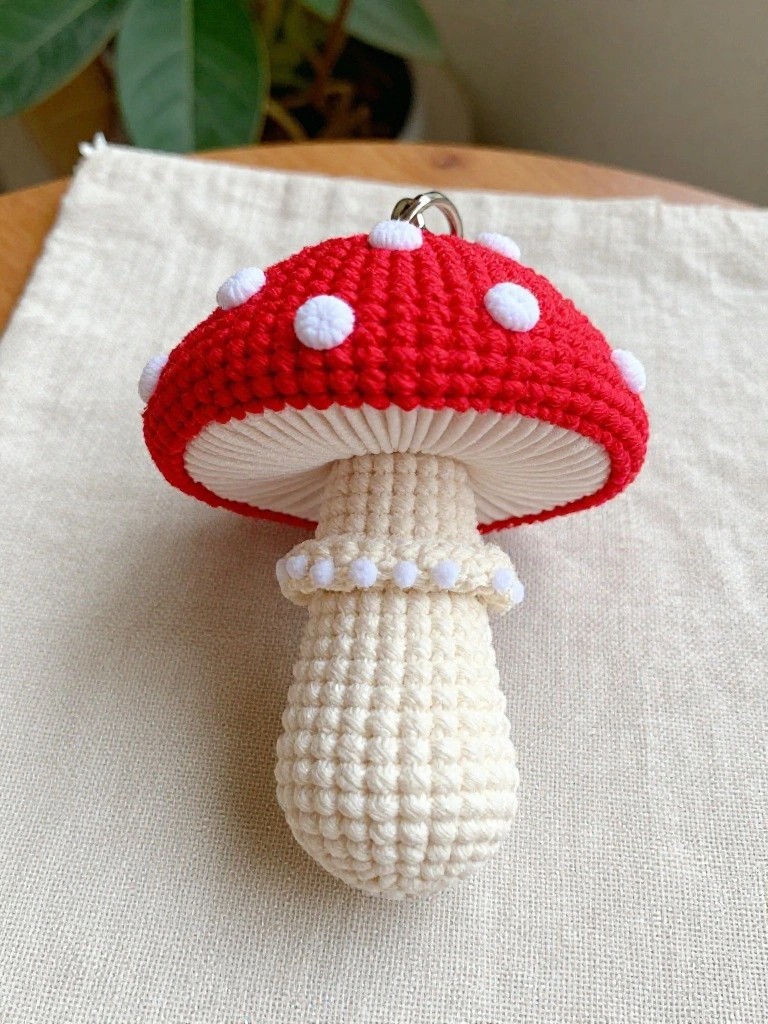 Crocheted red mushroom keychain with white spots on the cap and textured white stem, dangling from a metal keyring on a wooden surface.