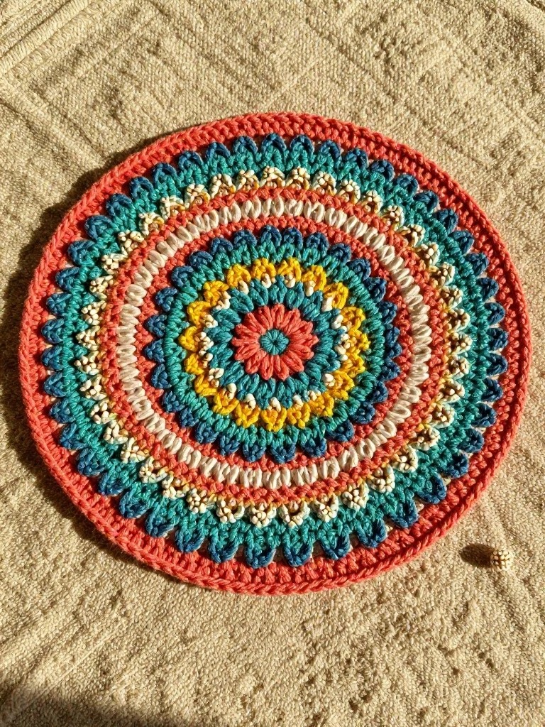 Handmade round crocheted mandala coaster in layered orange, turquoise, yellow, green, coral, and white on a beige woven background with a tiny seed for scale.