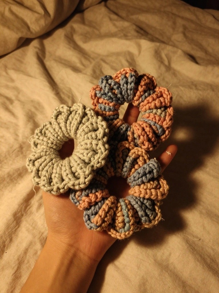 A hand displays three handmade crochet scrunchies, including a sage green petal-edged one and two striped in blue, peach, and coral, on a neutral bed.