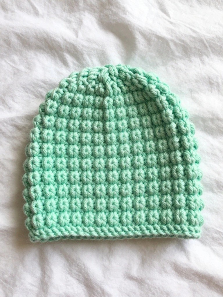 A small mint green crochet beanie with bumpy bobble texture across the crown, featuring a smooth bottom band, placed on white fabric.