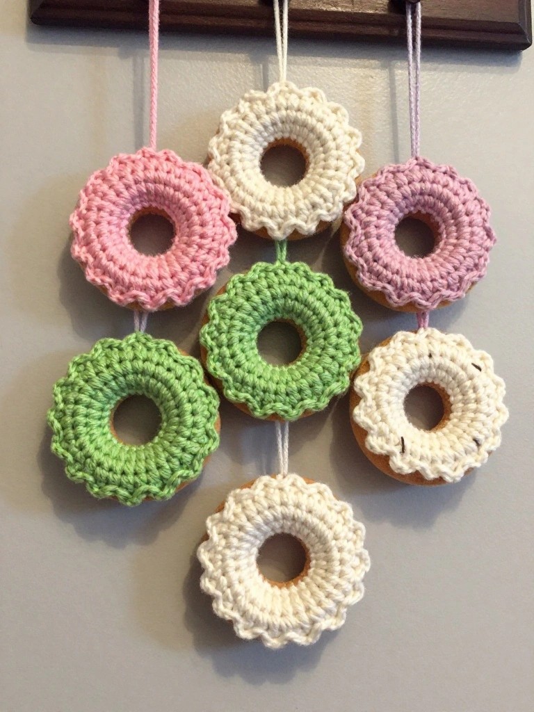 Wooden wall hanger displaying crocheted donuts in pink, green, purple, white, and beige, hanging by pink strings with textured stitches and icing-like details.