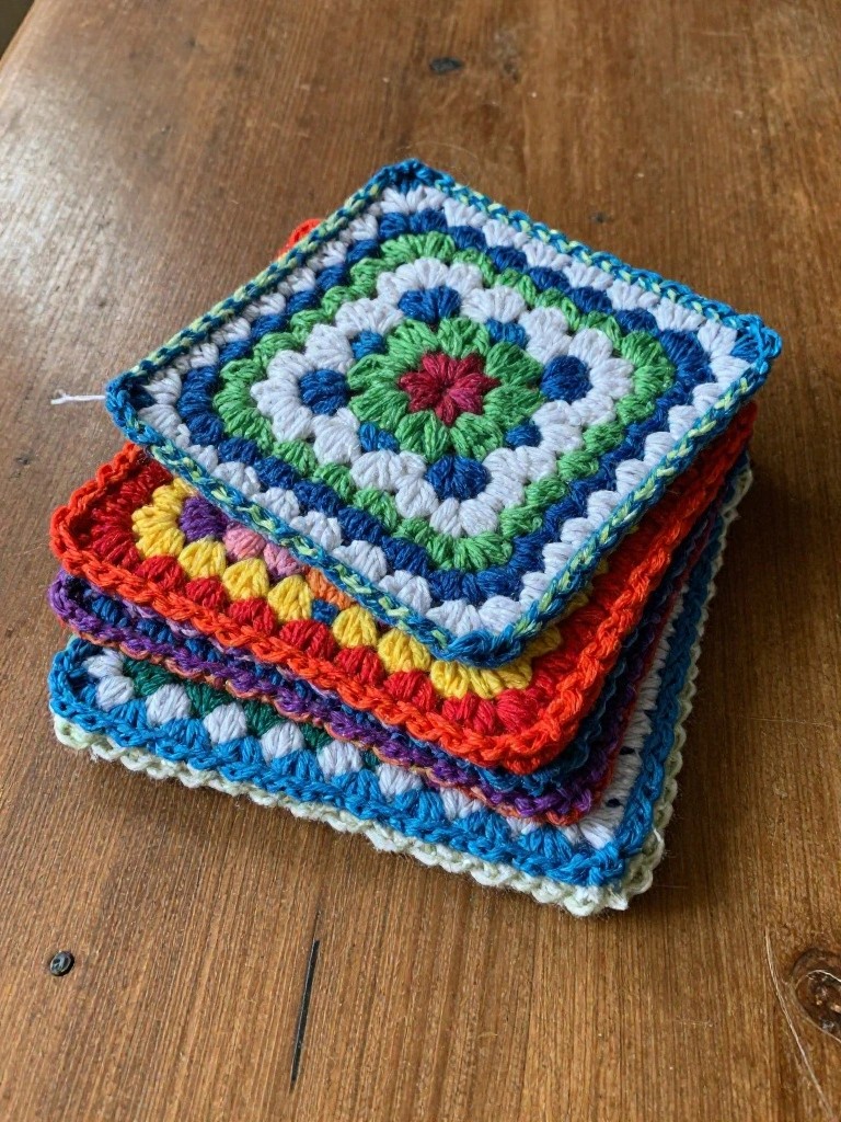 Stack of multicolored crochet granny square coasters on a wooden table, showing vibrant teal, green, red, yellow, orange, and purple shades with textured stitches.