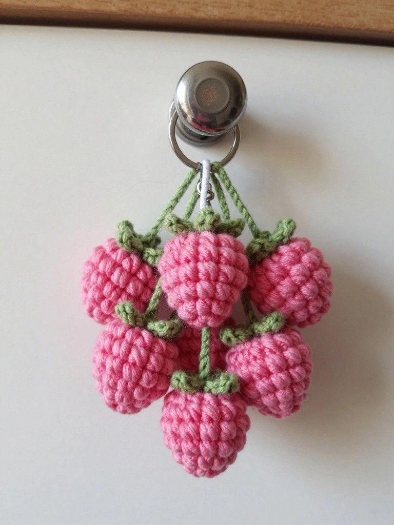 Close-up of a silver keyring holding a cluster of six tiny pink crocheted strawberries with green tops, dangling against a white drawer front.