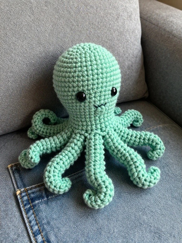Handmade mint green crocheted octopus plush with big black eyes, shy mouth, and eight curly tentacles sitting on a gray couch.