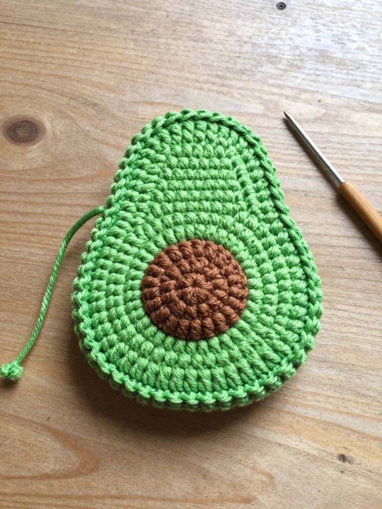 Crocheted green avocado-shaped pouch with brown pit detail and green cord loop sits on a wooden table beside a crochet hook.