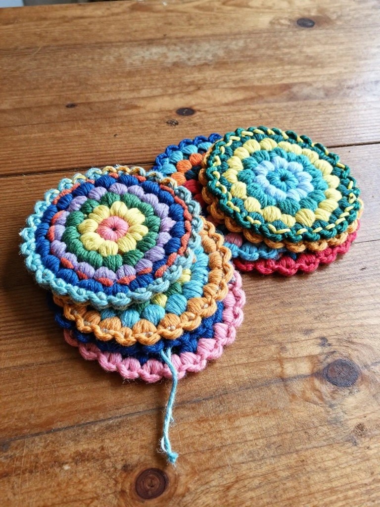 A pile of multicolored crocheted mandala coasters with scalloped edges stacked beside singles on a wooden table.