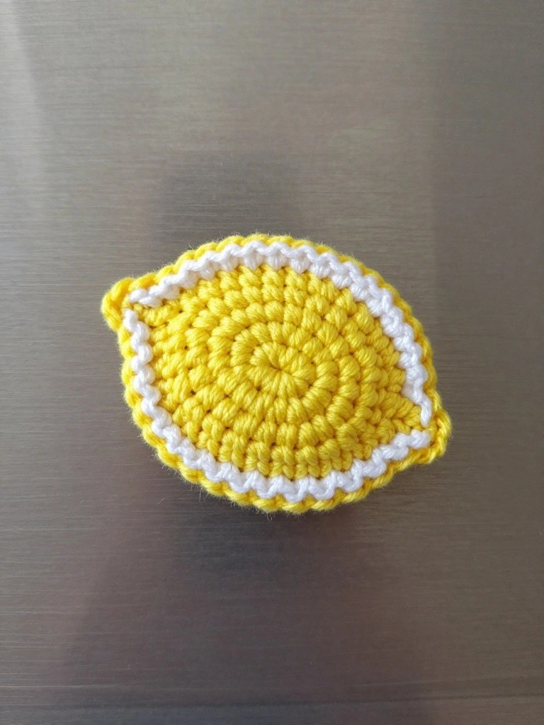 A handmade crocheted lemon slice in yellow yarn with white edging sticks to a stainless steel fridge door as a magnet.