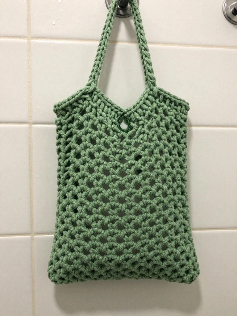 Handmade sage green crochet mesh tote bag with twisted handles hanging on a hook against white bathroom tiles.