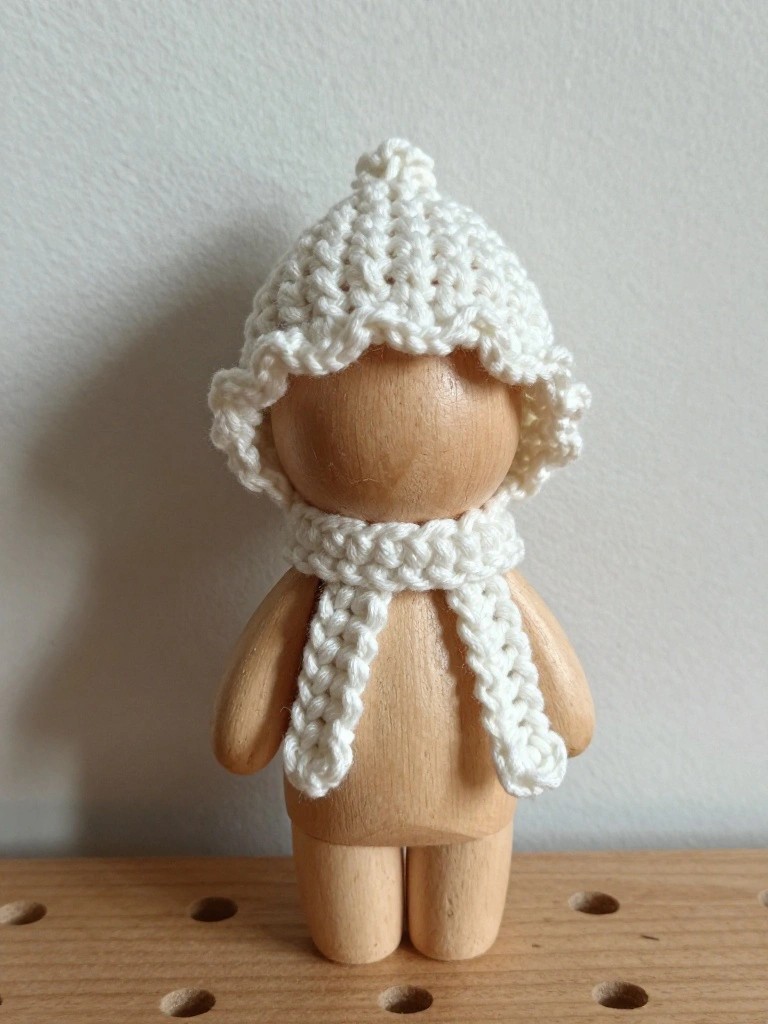 Small wooden doll with rounded unpainted body wearing a white crocheted hat topped with a pom-pom and ruffled brim, plus a matching wrapped scarf, posed on a pegboard surface.