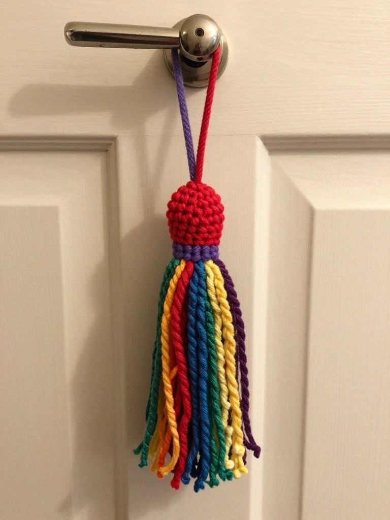 Crocheted red-headed tassel with rainbow yarn fringes looped over a silver door knob on a white door