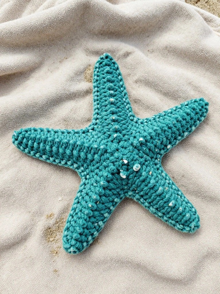 Crocheted teal starfish on a sandy beige fabric background with scattered sand.