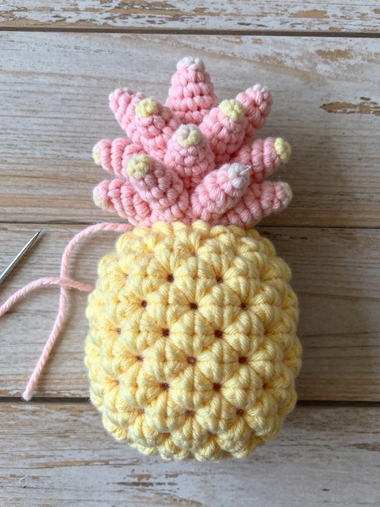 A partially finished crocheted pineapple with yellow body and pink top sits on a wooden surface beside a crochet hook and pink yarn strand.