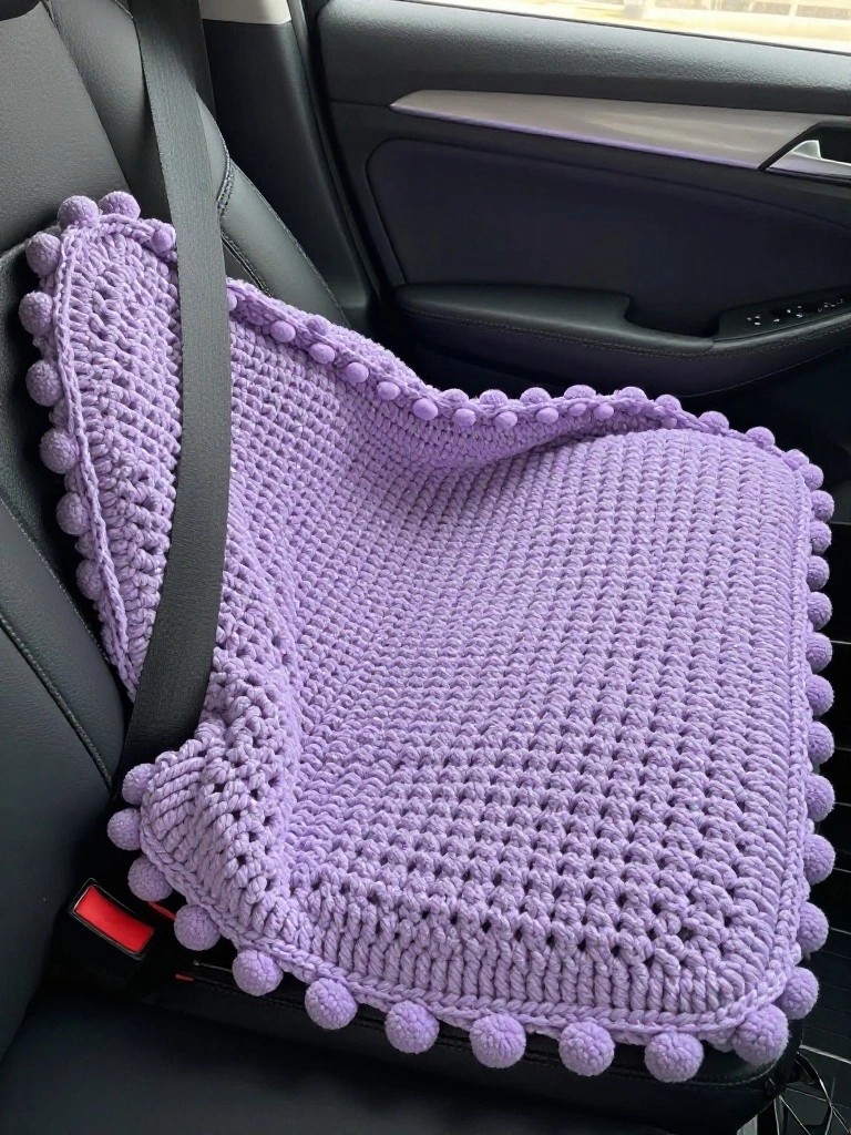 Purple crocheted blanket with pom-poms on a black car seat.