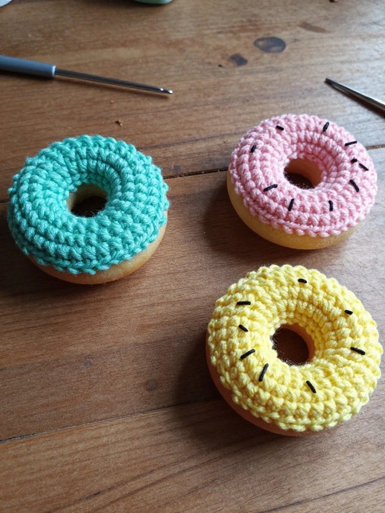 Three crocheted donut rings in turquoise, pink, and yellow rest on a wooden table beside two crochet hooks.
