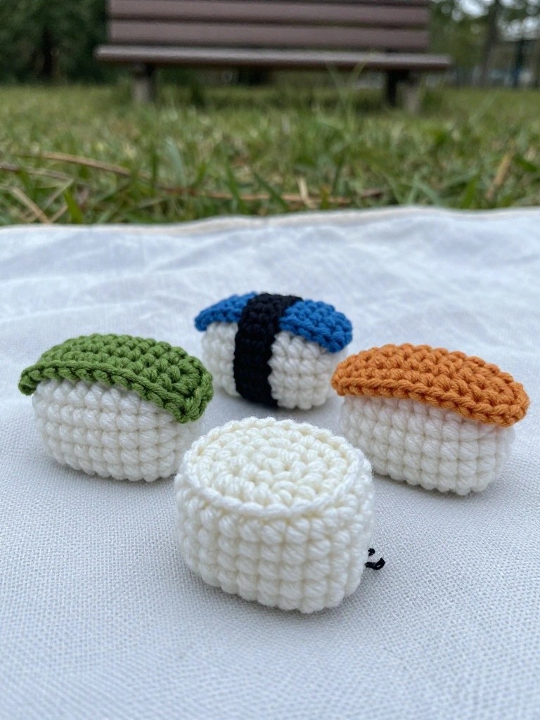 Four small crocheted sushi pieces sit on white fabric outdoors, showing green, blue-black, orange, and plain white toppings.