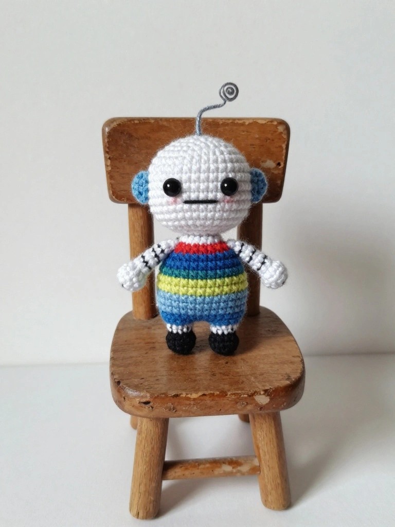 Crocheted white robot with rainbow stripes and antenna on wooden chair.