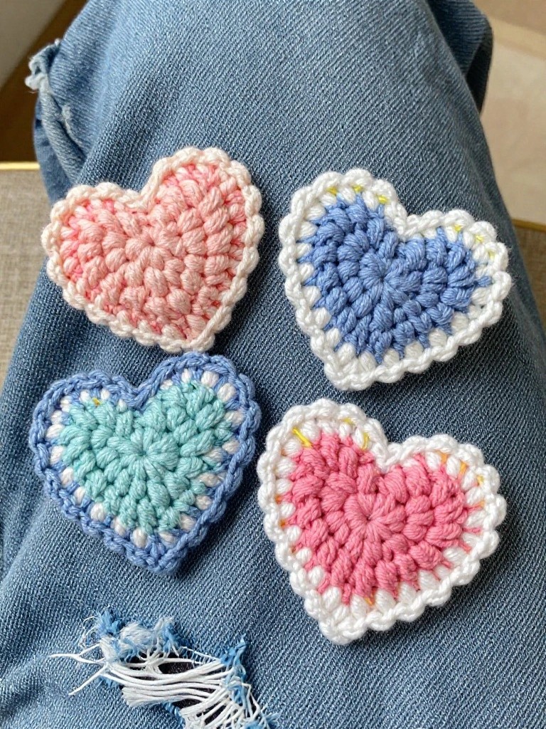 Four small crochet heart shapes in pastel pink, blue, and mint with white borders rest on blue denim fabric.