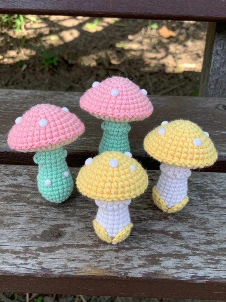 Four colorful crocheted mushrooms with spotted caps on a wooden bench.