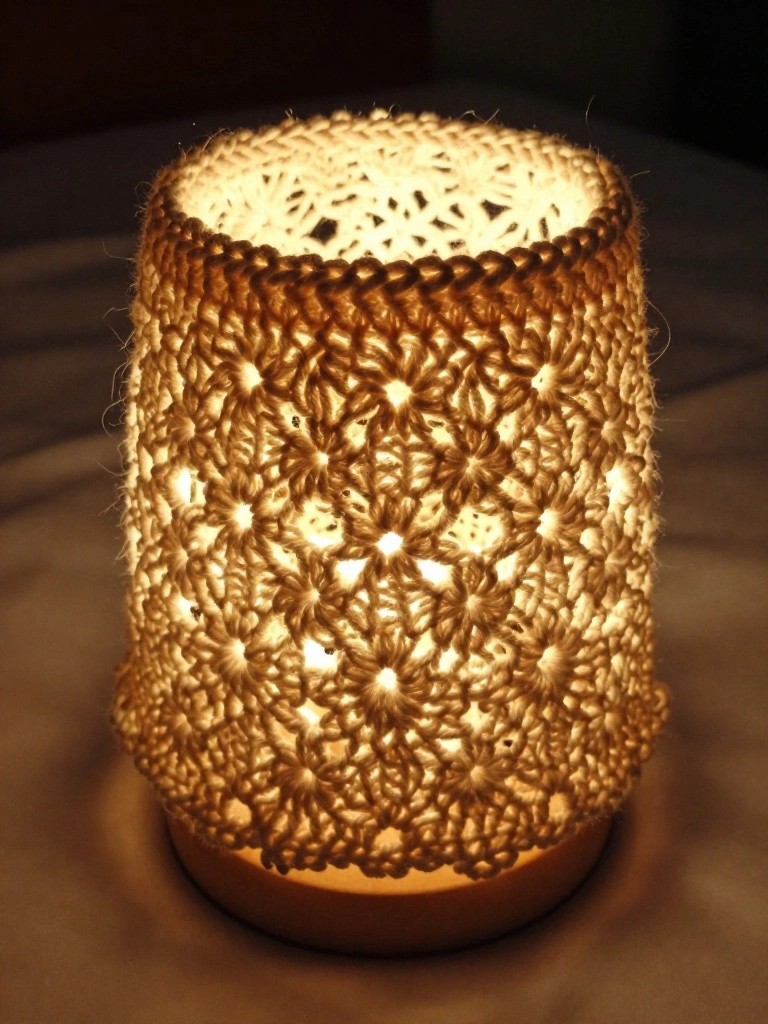 A beige crocheted cylindrical cover with open floral motifs sits over a glowing light base on a wooden surface.