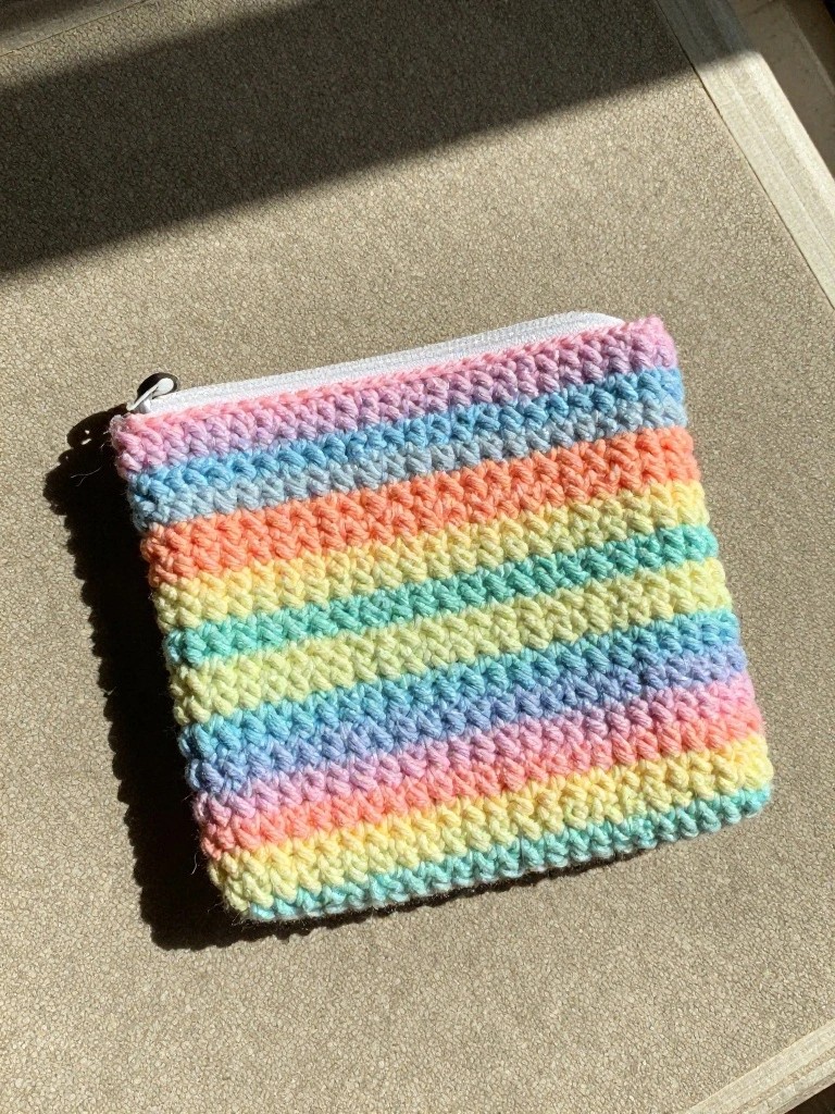 Crocheted rainbow-striped pouch with white zipper on sunlit textured surface