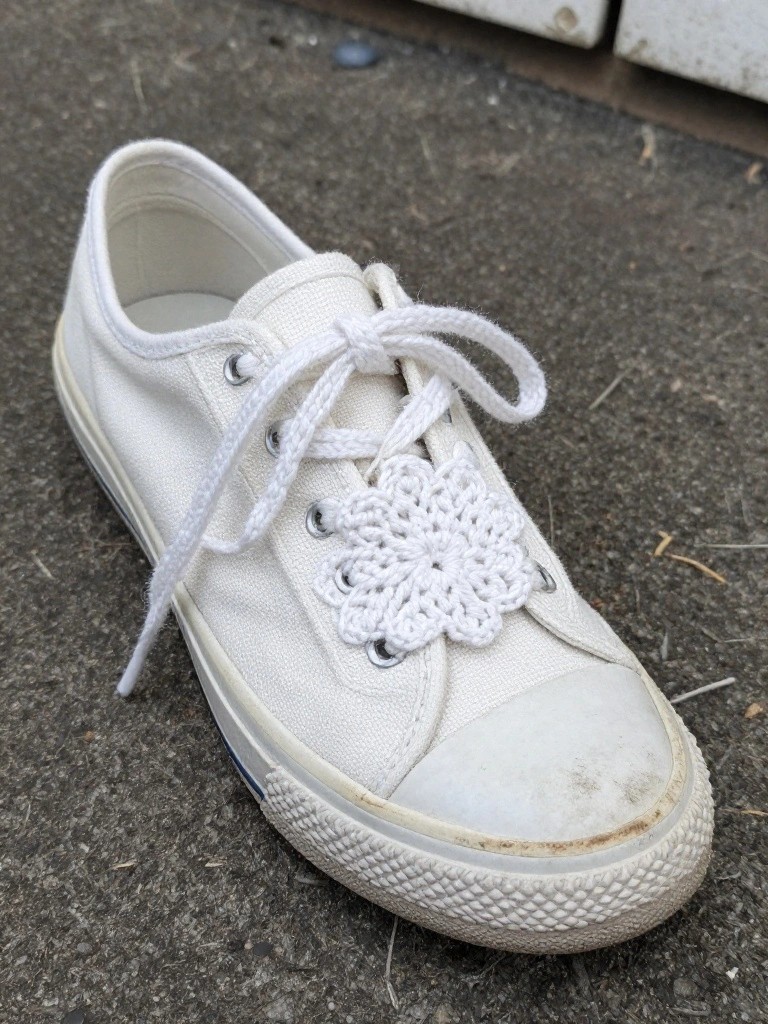 A white canvas sneaker with white laces and a small white crocheted flower resting on the laces.