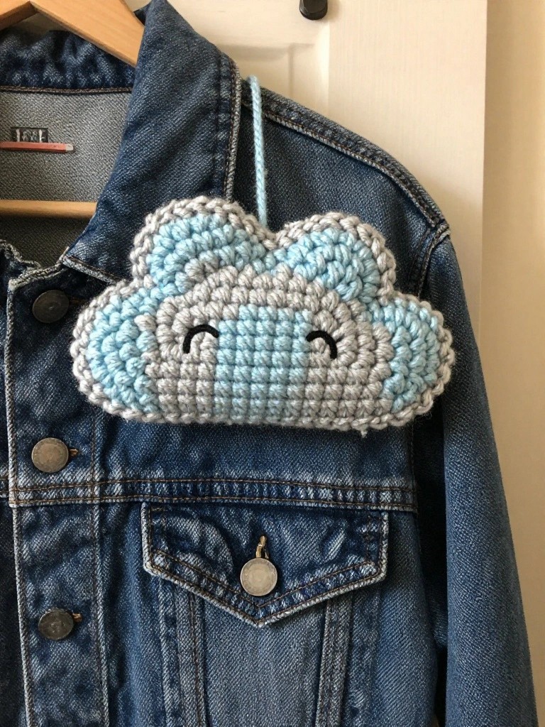 Crocheted light blue and gray cloud with stitched eyes attached by a cord to a blue denim jacket.