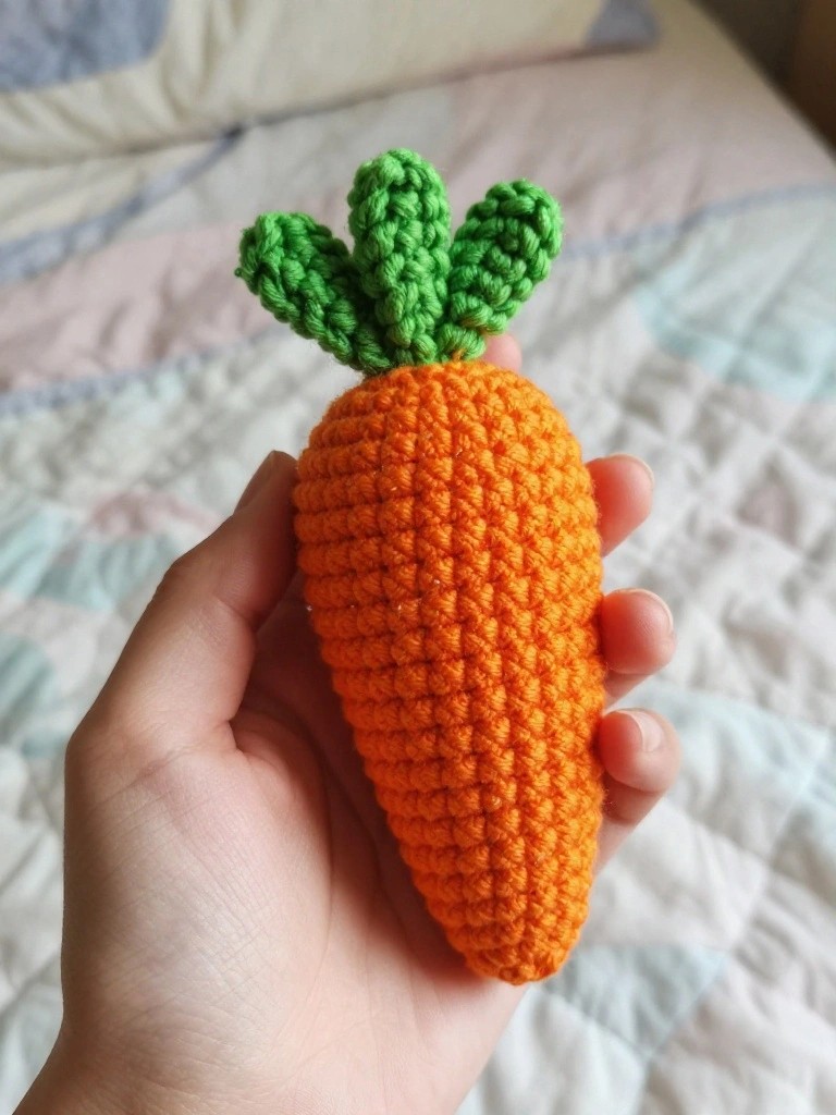 A hand holding a small crocheted orange carrot with green leaves on top.