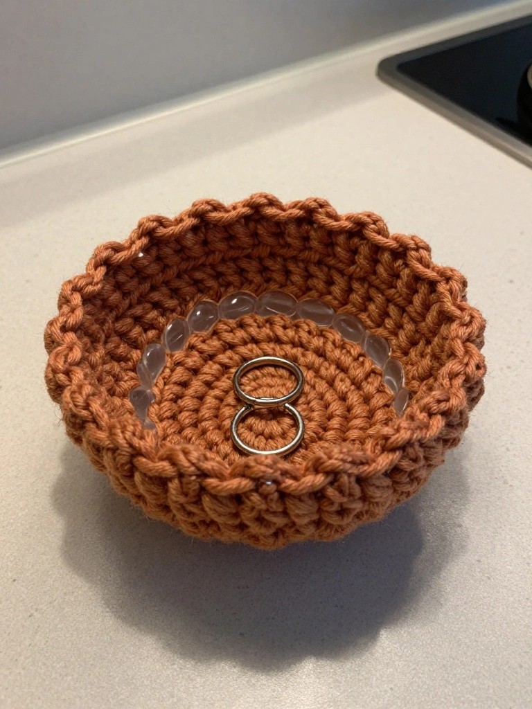 Rust-orange crocheted bowl holding interlocked silver rings and clear beads on countertop.