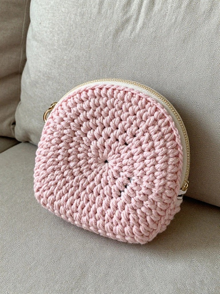 A small round pink crocheted pouch with a gold zipper and ring attachment rests on a fabric surface.