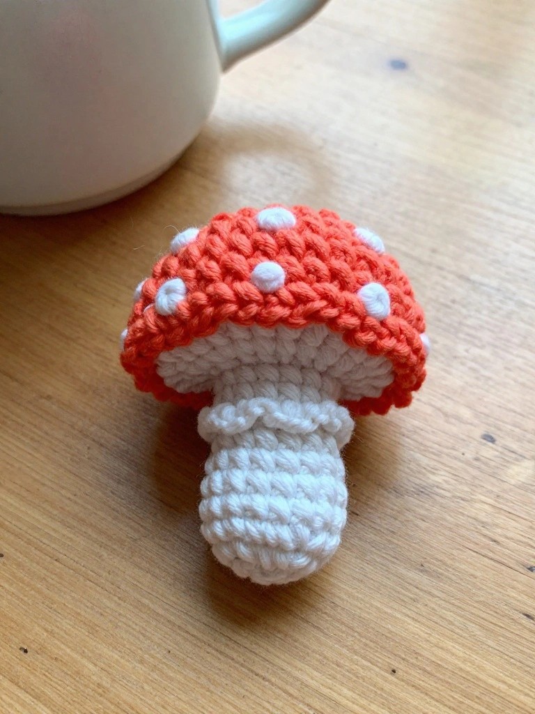A small crocheted mushroom with an orange-red cap dotted in white sits on a wooden table beside a white mug.
