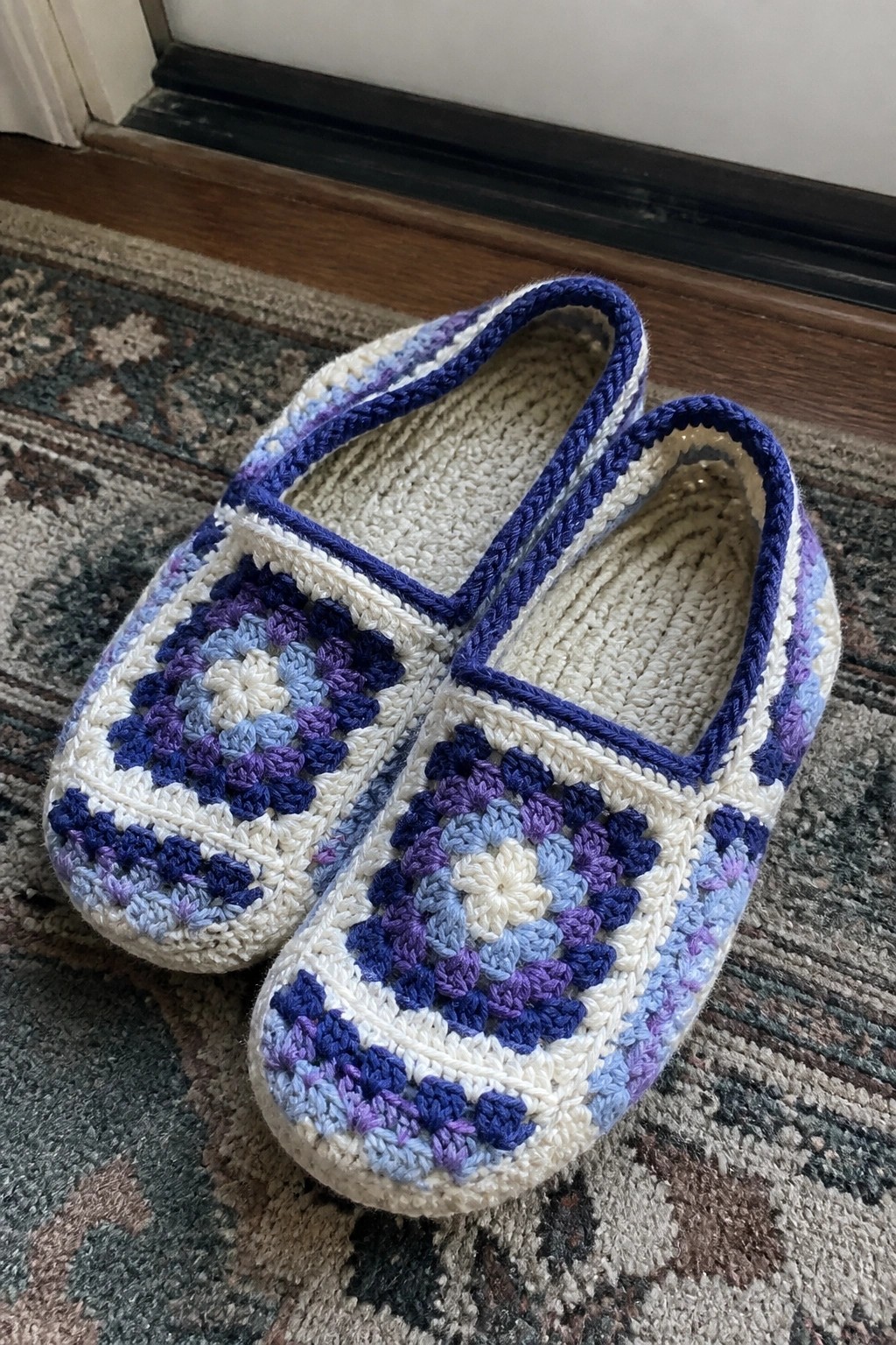 Pair of blue-purple crocheted granny square slippers on patterned rug.