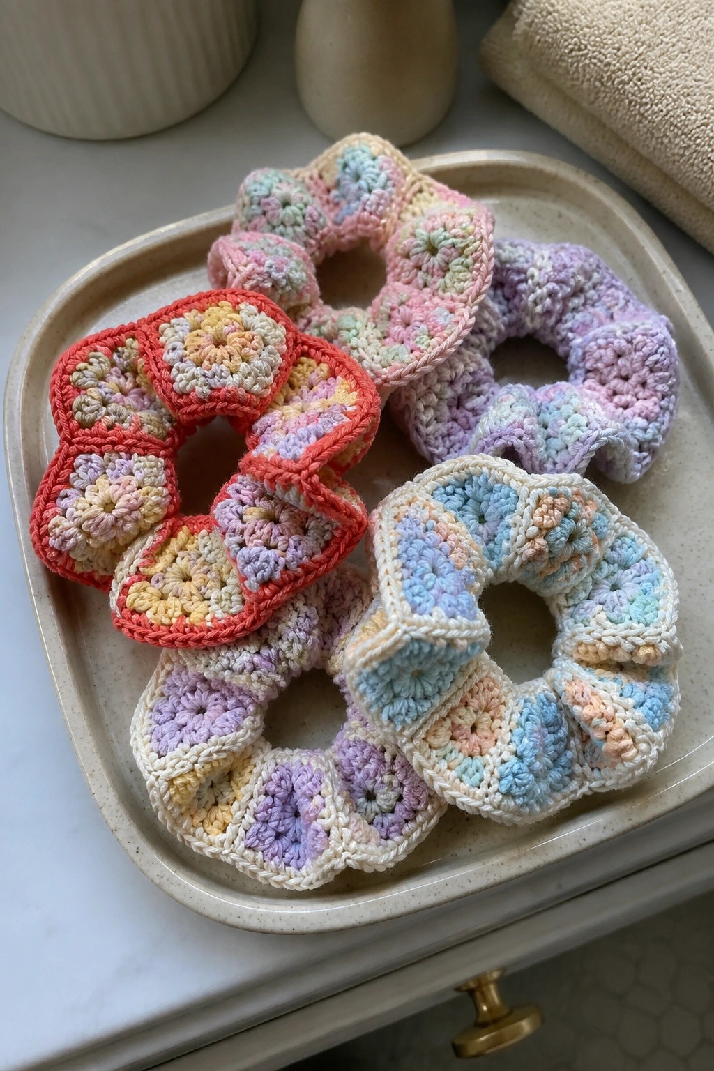 Several pastel crocheted granny square scrunchies on a beige tray.