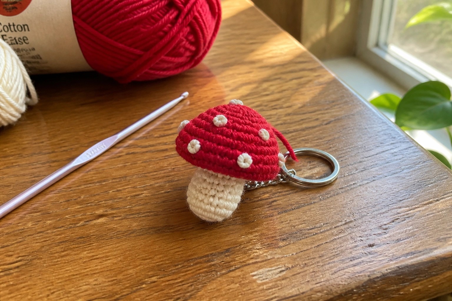 Red crocheted mushroom keychain with white spots beside pink hook and yarn balls