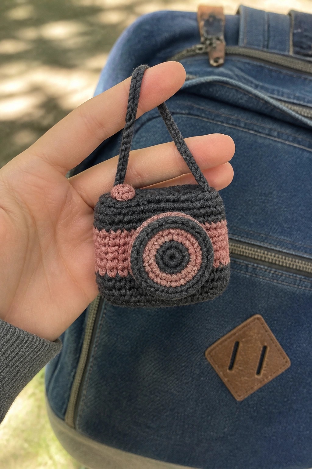 A hand holds a small crocheted camera keychain in gray and pink yarn with a braided strap.