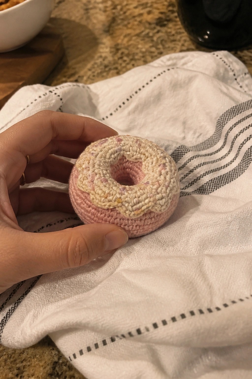 A hand holds a small pink crocheted donut with a cream top layer over a white cloth.