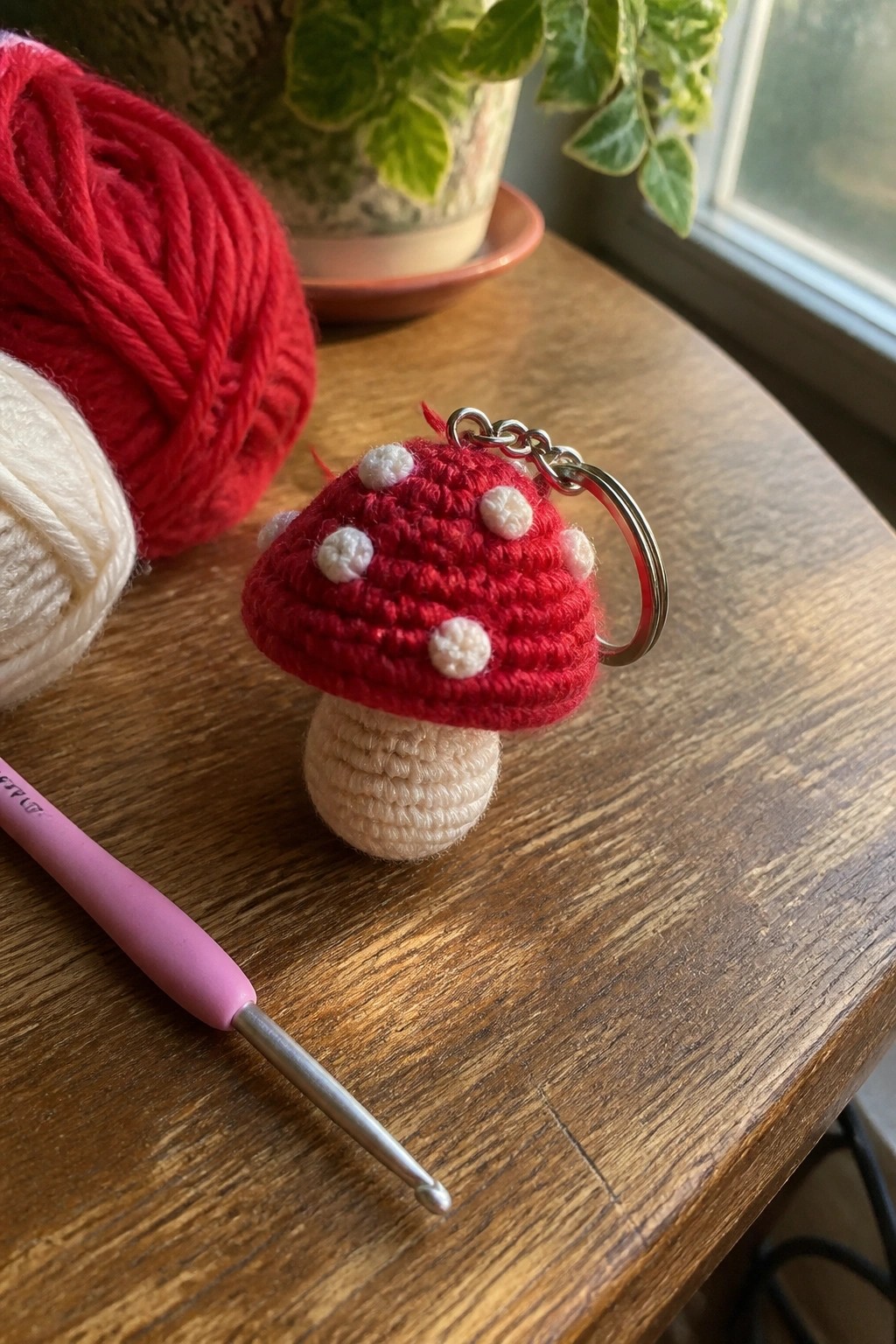 Red crocheted mushroom keychain with white spots beside pink hook and yarn balls