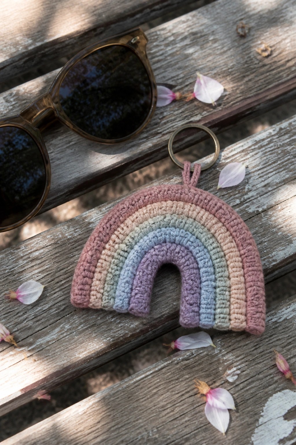 Pastel crocheted rainbow keychain beside sunglasses on wooden planks with petals