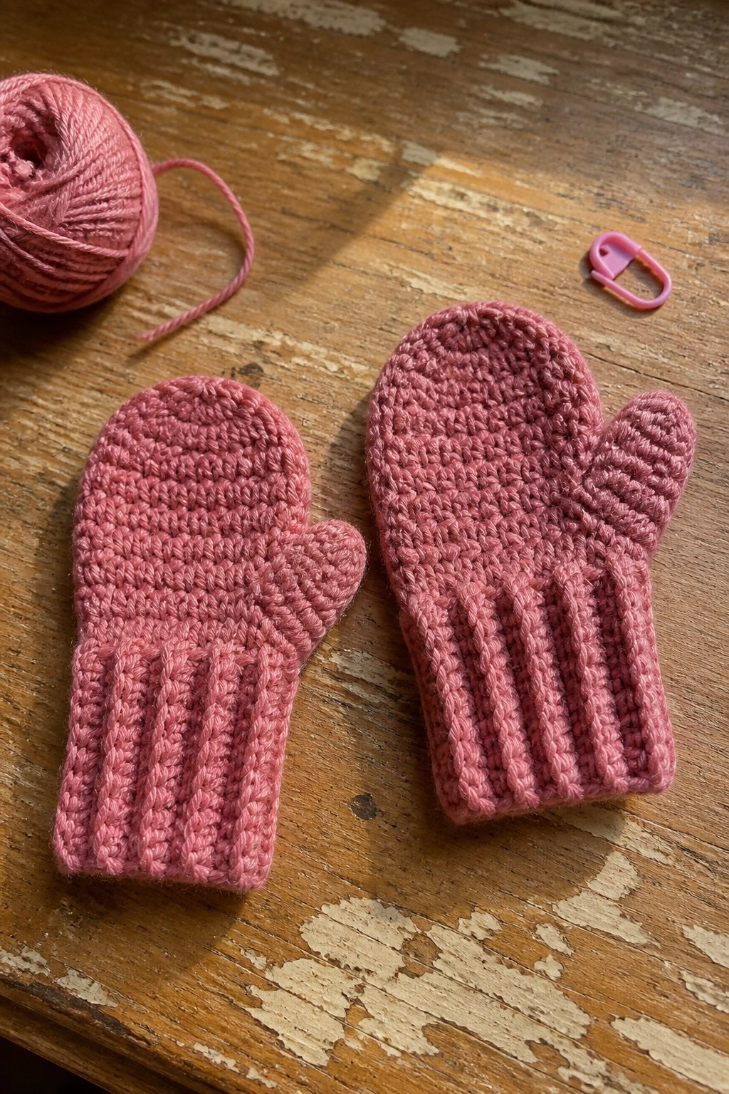 Two small pink crocheted mittens next to a ball of pink yarn and a stitch marker on a wooden table.