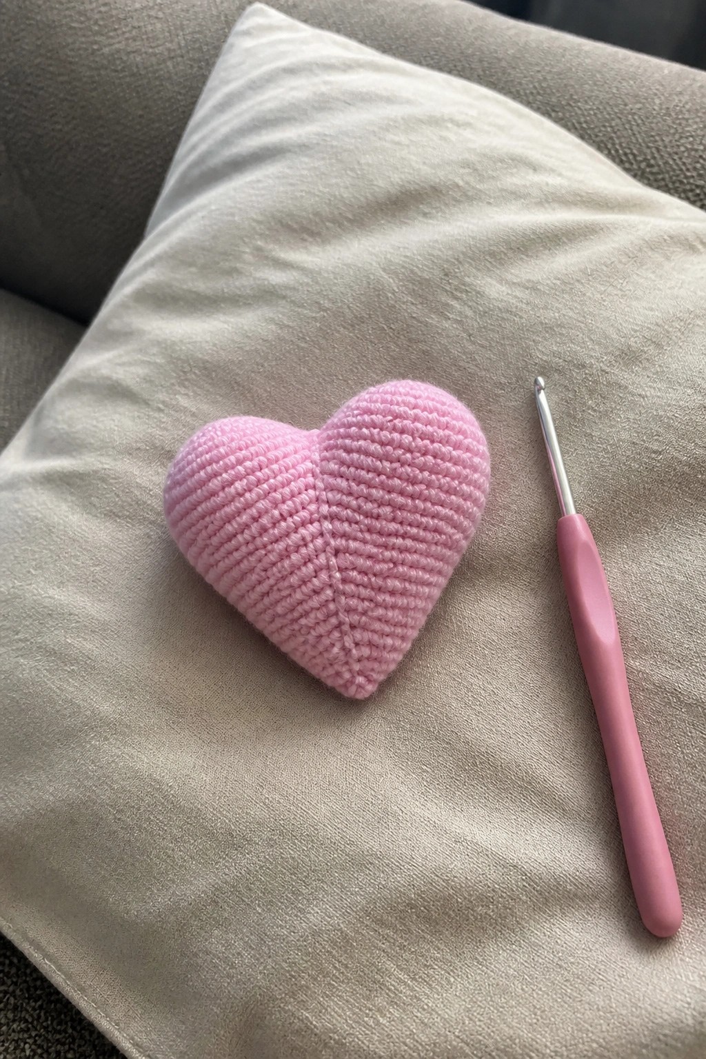 Pink crocheted heart beside pink-handled crochet hook on beige fabric cushion