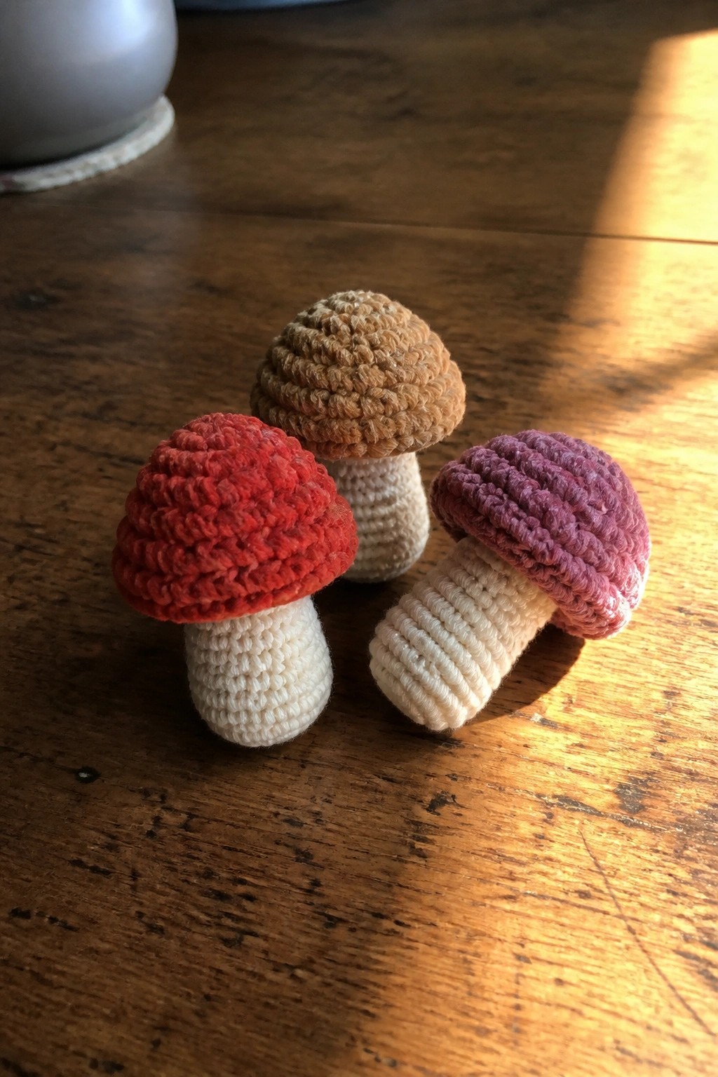Three crocheted mushrooms with red, brown, and purple caps on wooden table.