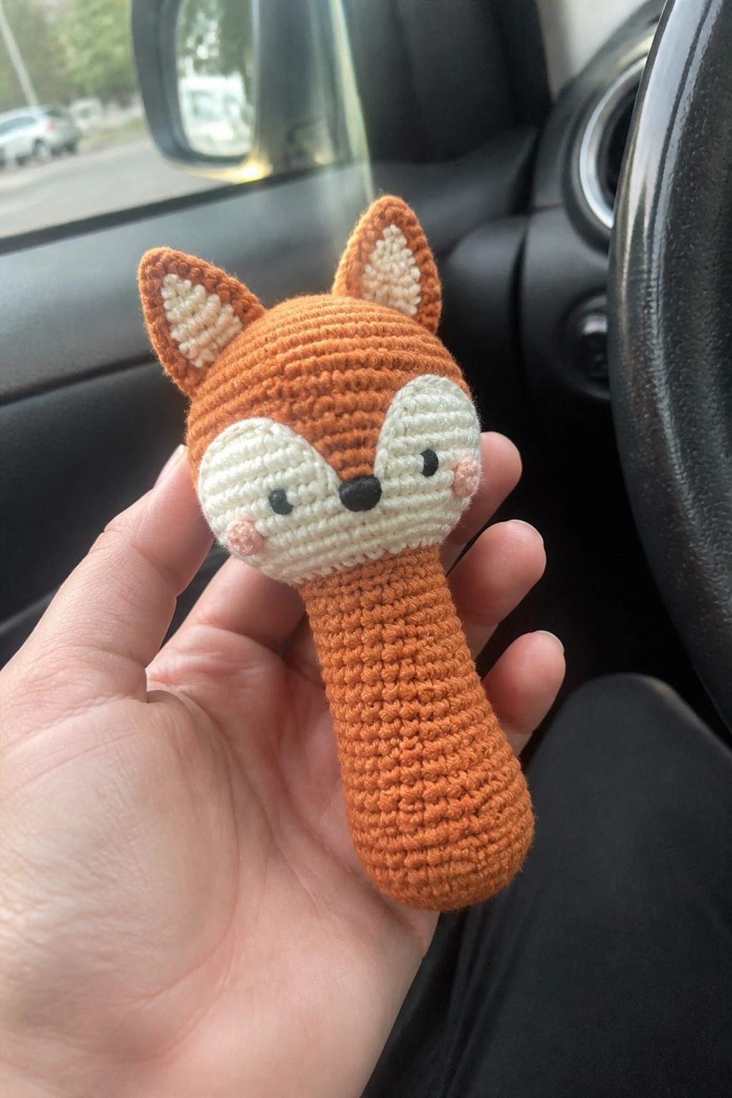 Hand holding crocheted orange fox toy with white face in car interior.