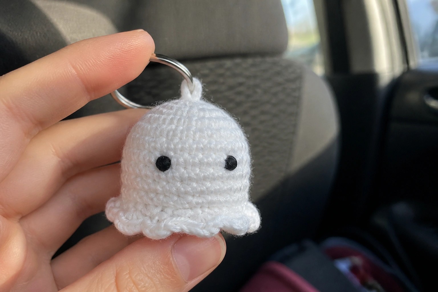 Hand holding tiny white crocheted ghost keychain with black eyes and metal ring.