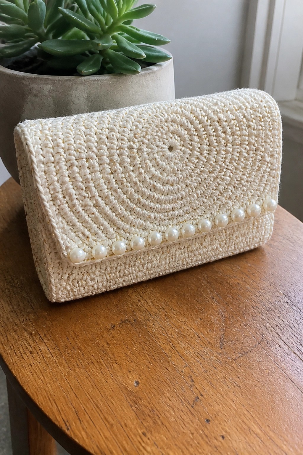 White crocheted clutch purse with pearls on wooden table beside succulent plant.