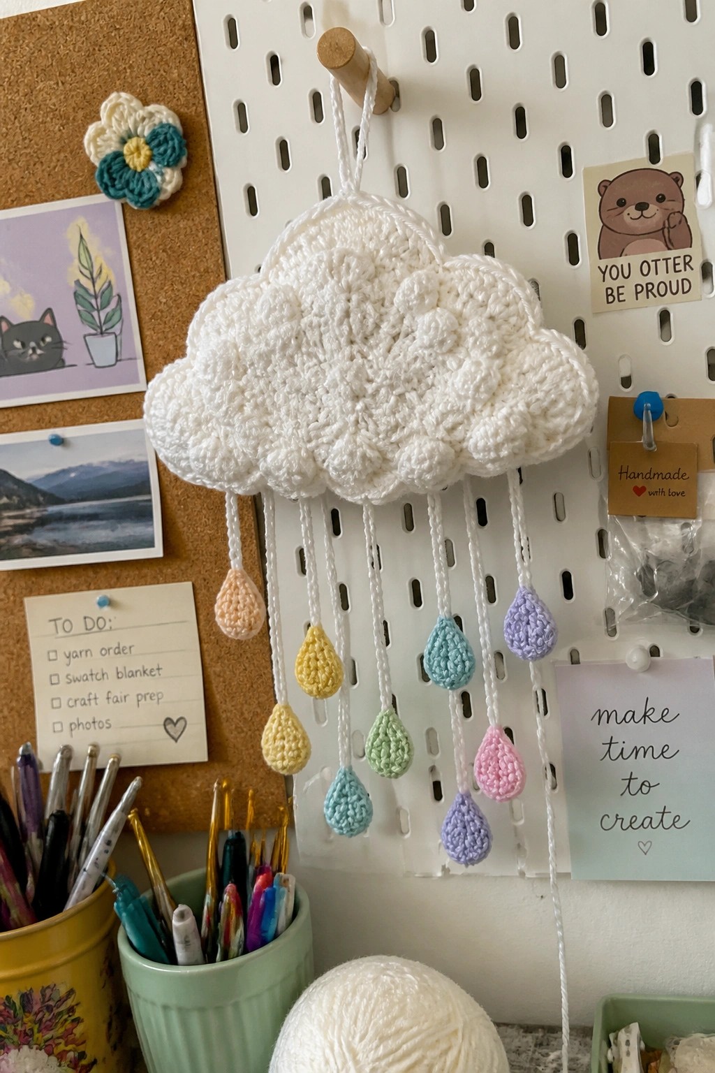 A white crocheted cloud with pastel teardrop shapes on strings hangs from a wooden peg on a white pegboard.