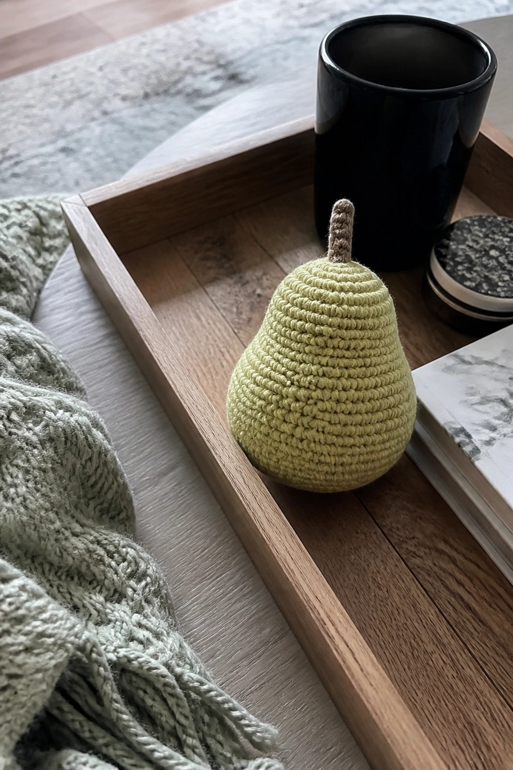 Green crocheted pear on wooden tray beside black cup and knitted blanket.